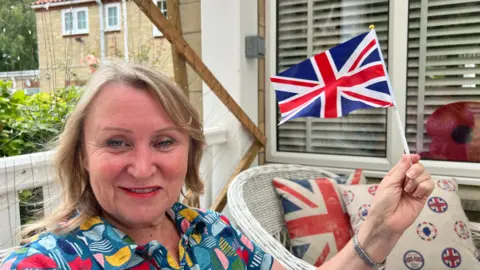 BBC A woman - Kim - wearing a brightly-coloured patterned blouse, holding a union jack flag in her hand. He has short blonde hair and is sat on a patio in a garden. A white wicker chair with union jack cushions can be seen in the background below a window with a cardboard poppy stood against it. 