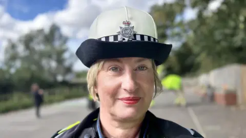 BBC News A female police officer with pale skin and blonde hair tied back. She is wearing a police hat with a Humberside Police crest and has red lips and red blusher on. The background of the picture is out of focus, but you can subtly see she's in a car park.