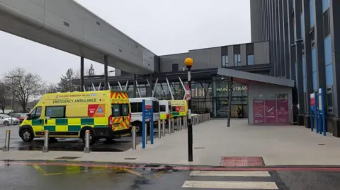 BBC Yellow and white ambulances parked outside the concourse of a large hospital building with a grey and blue tower block to the right, a modern glass-fronted atrium in the centre, and an enclosed grey walkway overhead.