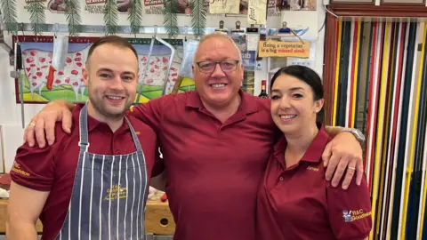 Richard Madden/BBC Three people in maroon shirts stand together in a butcher shop, with one wearing a striped apron. Their arms are around each other. Behind them is a selection of meat on a chopping block alongside knives and saws. A door on the right of the image is obscured by colourful ribbons.