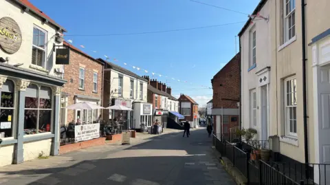 Richard Madden/BBC A sunny street lined with buildings including shops and cafes. On the left, a cafe has outdoor seating under white umbrellas. Colourful bunting has been strung across the street between buildings. A few people are walking along the pavement and in the distance. The sky is clear and blue.