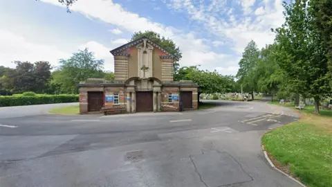 Google The chapel in Hull's Northern Cemetery. It is a redbrick and light stone building with a pitched roof and three large wooden doors at the front. It is surrounded by a road with trees and grass alongside and gravestones in the background.