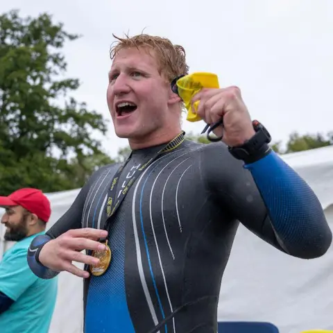 London Marathon Events Angus Leckonby in a blue wetsuit with short hair with a medal around his neck. There is a man to the side of him with a blue T-shirt, red cap and beard. There are some trees.