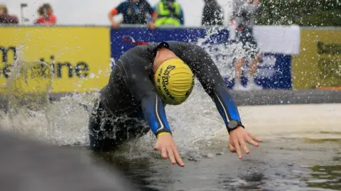 London Marathon Events Man in blue wetsuit with yellow swim cap diving into open water. There are people watching on in the background and trees. There is a yellow and blue Swim Serpentine banner. 