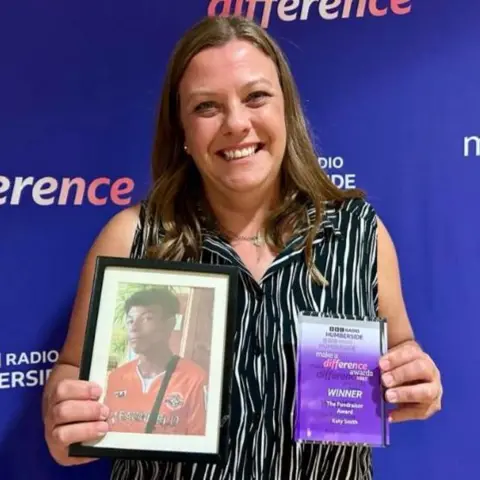 Charlie O'Loughlin/BBC Katy Smith, a woman with shoulder length, brown hair, is holding her Make A Difference award in her left hand. In her right hand, she is holding a framed photograph of her late son Ruben who is wearing a red sports top. 