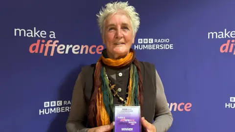 A woman with short, cropped blonde hair smiles at the camera. She is in front of a purple back drop reading: 'Make a Difference' and 'BBC Radio Humberside'. She wears a gold, brown and green striped scarf and had a brown waistcoat and long sleeved top underneath. In her hands is a purple Make a Difference Award reading 'Winner'.