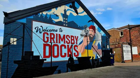 Creative Start Arts In Health CIC A large blue painting displayed on a brick wall. The mural details the words: "Welcome to Grimsby Docks" in bold black and red writing. Alongside the words- there is a bearded fisherman holding a large fish to the right. There are also silhouettes of boats and trawlers at the bottom of the wall. The mural is next to a red bricked building with a white and black poster on- the words are illegible. 