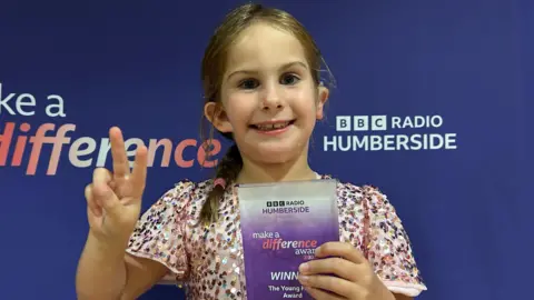Charlie O'Loughlin/BBC A young girl is seen smiling directly at the camera in front of a purple back drop. She is holding up two fingers in a peace sign, and is holding a BBC Radio Humberside Make a Difference Award for The Young Hero category. She is wearing a pink glittery dress, and wears her hair in a side plait. There is a cochlear implant on her right ear.