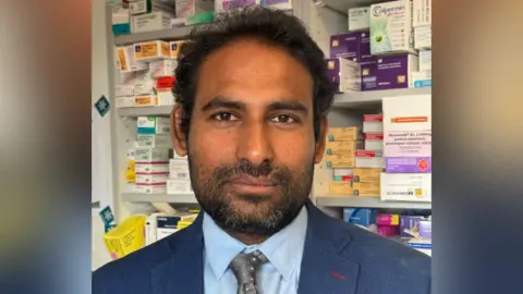 Amar Garlapati A head and shoulders shot of a man with black hair and black stubble and a moustache. He is wearing a blue suit and shirt with a grey tie with dots on it. Behind him you can see shelves full of different coloured medications. 