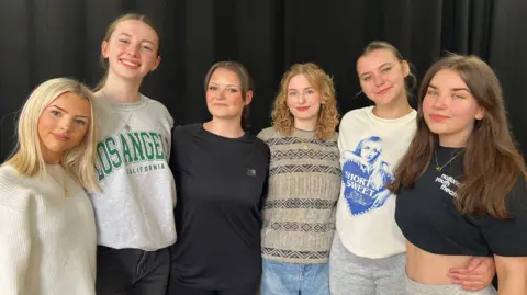 BBC Six young women stand arm-in-arm facing the camera in front of a black curtain. All are smiling and wearing casual clothes. 