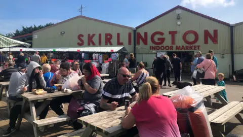 Skirlington Market People sit around wooden picnic tables, some eating food, outdoors at a market site. In the background is a warehouse-type building with a sign reading "Skirlington" in huge red letters on a cream-coloured wall, and "Indoor Market", in smaller red letters. Market stalls can be seen in front of the building with a variety of clothes hung up.