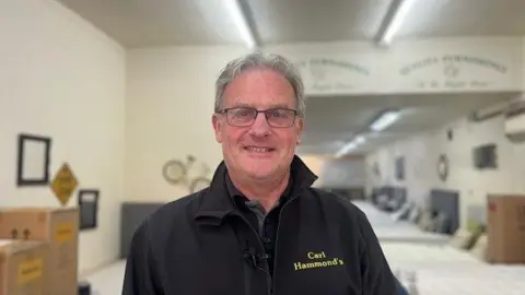 Man wearing glasses and a black fleece which says Carl Hammond's on it. He is standing in the furniture store which he owns. In the background are beds which are for sale.