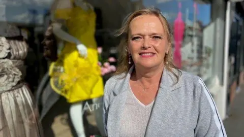 Diane is standing outside her shop, which has mannequins in dresses in the window. She is wearing a grey blazer and a white v-neck top and has long blonde hair. She is wearing pink lipstick and dangly earrings and is smiling at the camera. 
