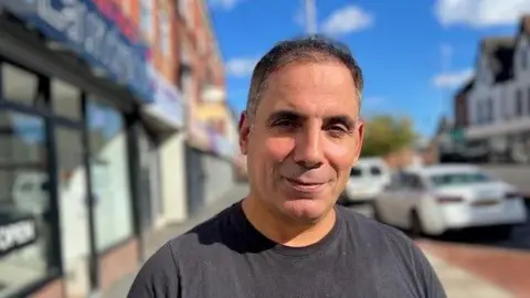 Man with short dark hair wearing a grey t-shirt standing on a busy roadside with shops either side and parked cars.