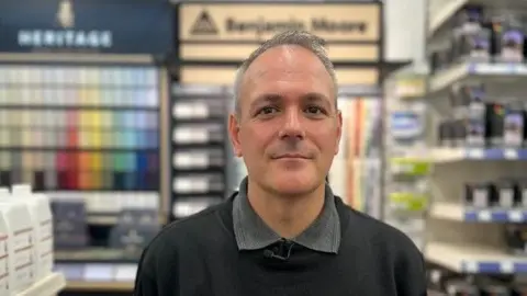 BBC Man with short hair wearing a grey shirt and black jumper. He is a standing in a paint shop with numerous paints and samples stacked up on the shelves behind him. 