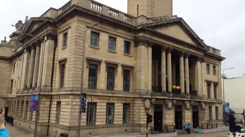 BBC The Guildhall is made of stone and has four large pillars that hold up an apex roof above a balcony on the first floor. It is a grand building with large black windows.