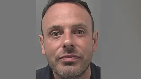 Humberside Police A police headshot of Daniel Cotson. He has short brown hair and is wearing a black polo shirt. The wall behind him is grey. 