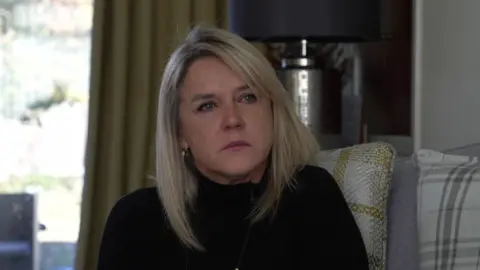 BBC Tearful Janet Skinner with shoulder length straight blonde hair wearing a black high neck jumper sitting on a grey sofa with checked cushions