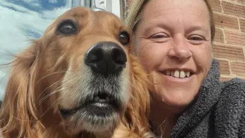 Katie Stevens Golden retriever and Irish retriever cross dog with a woman with blonde / brown hair smiling wearing a grey fleece. There is a window and a brick wall behind them.