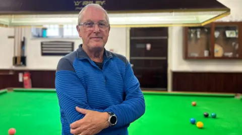 Charlie O'Loughlin/BBC A man looks directly towards the camera standing side on. He is stood in front of a large green snooker table with multicoloured snooker balls on top. He wears a blue pullover jumper with dark patches on the shoulder and neck. A large black smart watch is on his right arm.