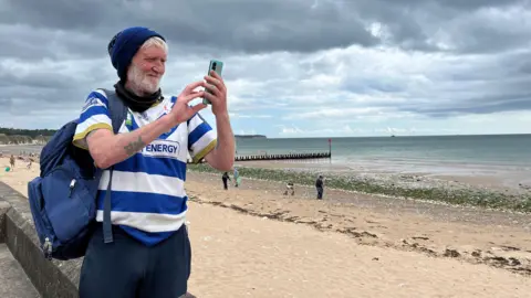 JESS LORD/BBC NEWS An older man is taking a photo of the beach. He is using an Android phone to do so. He is wearing navy tracksuit bottoms, a blue and white sports shirt (Halifax Panthers) and a navy blue woolly hat. He has a large dark blue rucksack on his back. The majority of the image is of the beach - sand, sea and a sky covered in white and grey clouds. The background is blurred but still visible - the seafront can be seen. 
