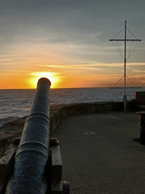 Kev Oldfield A photograph of a canon overlooking the sea, the placement of the sun is at the end of the canon. The sky is a mix of golden orange and dusty blues. The sea looks dark blue and calm.