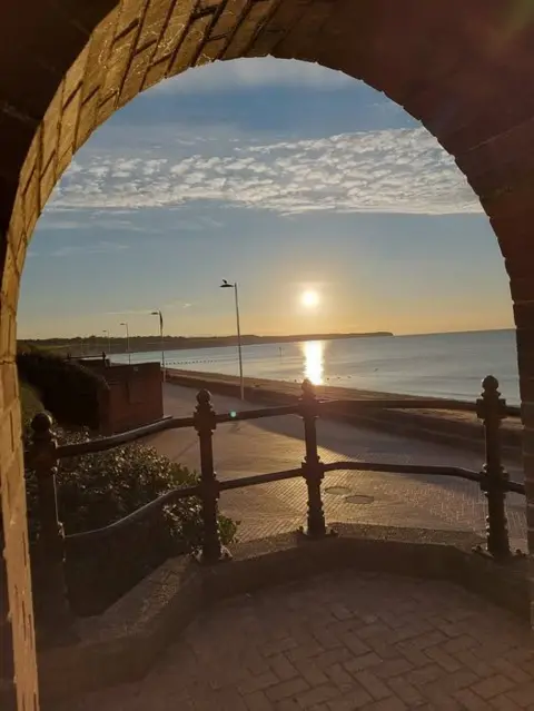 Kev Oldfield A photograph of the Bridlington seafront, taken behind a set of railings which are in shadow. The sun is rising. The sky is blue with hints of yellow and the sea is still and very blue. 
