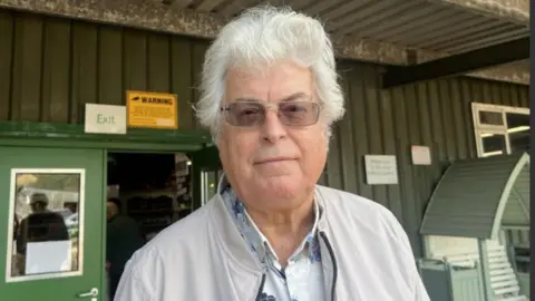 A man with short curly grey hair is wearing dark spectacles and a white and blue floral shirt under a cream baseball-style jacket.