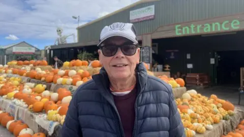 A man with blue jacket and dark red jumper wearing sunglasses and a black and white cap is standing in front of a collection of pumpkins at a garden centre and looking into the camera.