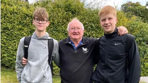The Market Weighton School A grandfather standing arm in arm with with two boys. One boy has a mop of brown hair and wears a grey hoody and black rucksack. The grandfather has white hair and wears a black zip top. The second boy has short ginger hair and wears a black tracksuit top and black rucksack. They are standing in front of green foliage.