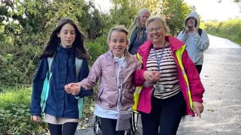 The Market Weighton School Three girls and two women walking down a country lane, with green trees and bushes to either side. In the front row, a girl has long brown hair, a blue jacket and black leggings. She is holding hands with a second girl who has blonde hair, a pink jacket and black leggings. Her other hand is held by a woman who has blonde hair, glasses, pink jacket, striped top, lanyard and dark trousers. In the back row a woman with long blonde hair and a dark top walks with a girl who has long blonde hair, a grey hoody and dark trousers.