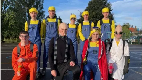 The Market Weighton School Nine students dressed in fancy dress, posing in two rows in a school playground. Those standing at the back have yellow tops, hats and blue dungarees. Those in the front are kneeling. One wears an orange outfit and glasses, another wears black clothes, a bald cap and a long nose. A third wears a king's robes and crown. A fourth wears a white gown and glasses. 