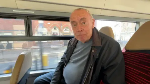 A man with short cropped grey hair sits on a bus seat. He's wearing a blue and grey bomber jacket and a grey t-shirt.
