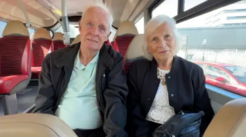 A man and woman sit next to each other on bus seats. Jim, on the left is wearing a black jacket and teal coloured shirt. Doreen on the right has a lace white top, black jacket and a black handbag. The both have grey hair.