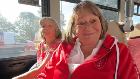 Two women sit next to each other on a bus. There is a white van driving by out of the window. Both women have neck length brown hair and are smiling. Susan, in front and on the right, is wearing a white blouse and a Hull Kingston Rovers red hoodies, which is open at the neck.