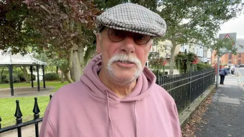 A man wearing a grey and brown checked cap, stands beside a black iron fence overlooking a green area with trees and a bandstand. He has sunglasses on, and grey moustache, and wears a pastel pink hoodie.