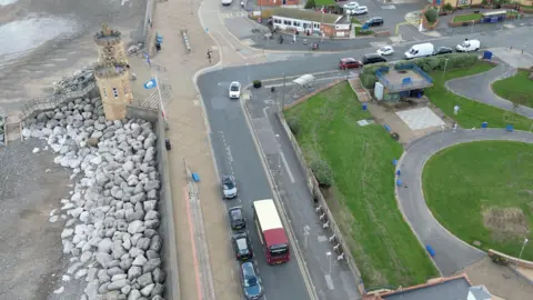 An aerial view of a yellow and red above passing by a park on one side of the road and approaching Withernsea's Pier Towers; two small yellowing towers with thin, narrow windows. The're surrounded by rocks and sand on oneside, and the pavement and road on the other. Steps lead from the towers down to the beach.