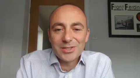 A man in a light blue open-necked checked shirt sits in front of glass door, with white walls and a picture on the wall. He has a receeding hairline and is smiling.