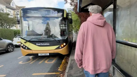 BBC A man in a pink hoodie, jeans and a checked flat cap prepares to board a single decker bus at a bus stop. The bus display says it is the 129 to Withernsea. The bus is yellow and black, and Ian has his back to the camera.