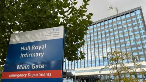 BBC A sign in the foreground reads Hull Royal Infirmary Main Gate. Behind, the tower block is visible. The frontage is made up of identical rows of windows with strips of light blue cladding running horizontally and dark grey frames running vertically up the full height of the building.