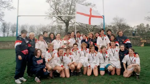 Getty Images The England team pose for a victory photo on a green field in front of white rugby posts. The 15 players are wearing white shirts and shorts with half standing in the back row and the rest kneeling in the front row. Coaches and support staff, wearing blue tracksuits, are also in the picture.