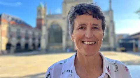 BBC News A woman smiles as she stands in a square in front of a large church. She has a short grey pixie haircut and wears a pale pink floral blouse. 