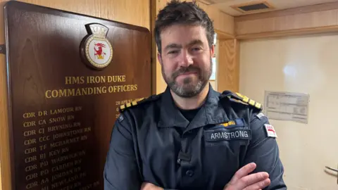 Dale Baxter/BBC The captain of HMS Iron Duke stands with his arms folded in front a wooden plaque displaying the names of former commanders. He is wearing a blue coloured naval uniform.