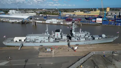 A grey coloured warship is moored at a dock. Shipping containers, cranes and industrial warehouses are visible in the background.