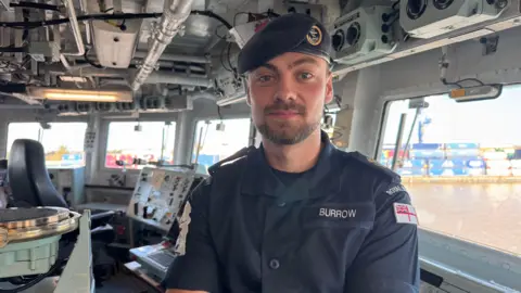 Dale Baxter/BBC A man wearing a naval uniform stands on the bridge of a warship. He is standing in front of an array of control panels with the surrounding dock area visible through the windows.