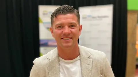 BBC Luke Campbell smiles at the camera after winning the mayoral election in Hull and East Yorkshire. He is wearing a beige blazer with a white T-shirt. He is standing in front of a stage that includes a podium and a black backdrop.