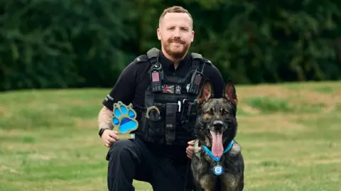 Thin Blue Paw Foundation PC Tom Marsden, who is wearing black police uniform and a police vest, kneeling on one knee with a blue pawprint trophy rested on it next to PD Yoiko, who is wearing a blue medal. PC Marsden has short brown hair and brown facial hair. PD Yoiko's ears are perked up and his tongue is hanging out of his mouth. They are in a grassy area with green bushes behind them. 