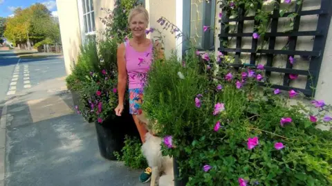 Submitted A woman with blonde hair and a full fringe tied back is wearing a bright pink vest and blue and pink shorts. She is smiling at the camera and standing next to a black and silver front door. Next to the front door are several purple flowers and greenery that she has planted. You can see the pathway in front of her and a beige-coloured dog is sat next to her, looking up at her.