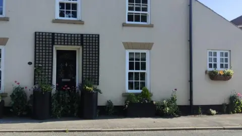 Judith Seeley The front of a beige house with a black front door at the side of a road. You can see two tall planters with flowers on each side of the front door, a shorter but longer one underneath a window and more greenery and flowers surrounding them. There is a footpath in front of them.