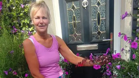 Submitted A woman with blonde hair and a full fringe tied back is wearing a bright pink vest and blue and pink shorts. She is smiling at the camera and standing next to a black and silver front door. Next to the front door are several purple flowers and greenery that she has planted.